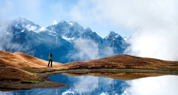 Une personne debout près d'un lac reflétant avec des montagnes enneigées et des nuages.