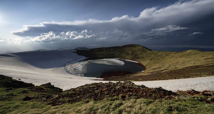 Lac de cratère et terrain enneigé sous un ciel dramatique.