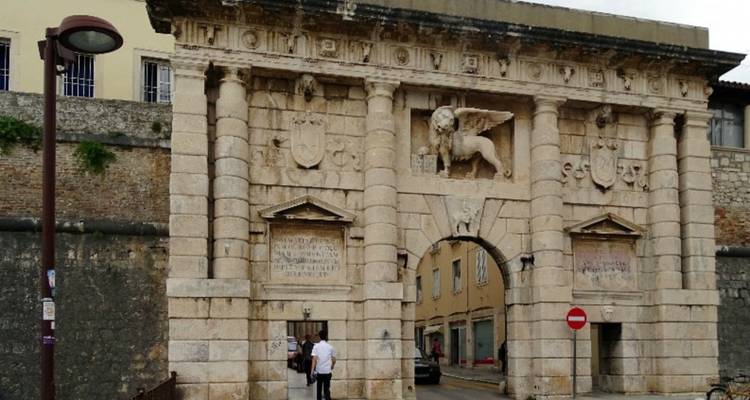 Historische stadspoort met sculptuur erboven, mensen die voorbijlopen.