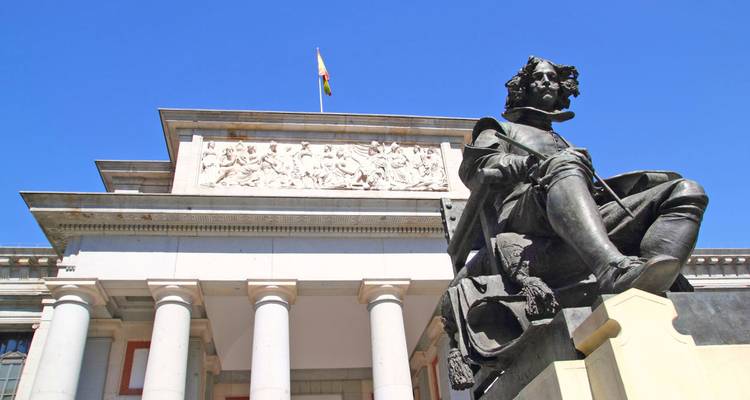 Statue in front of the Prado Museum building under a clear blue sky.