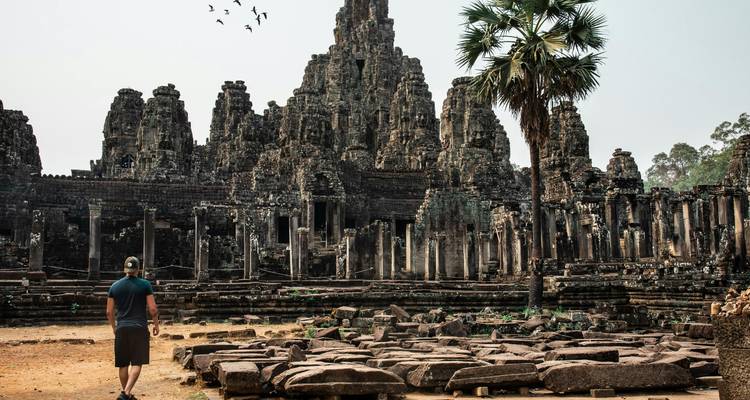 Un viajero solitario camina hacia las intrincadas torres de piedra de una ruina de templo jemer con pájaros volando por encima.