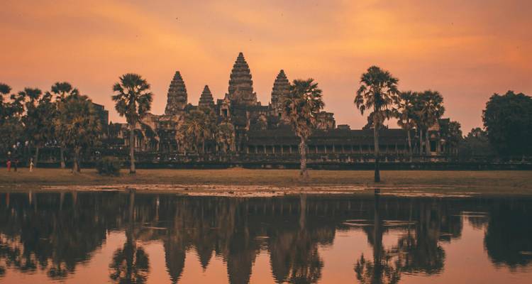 Angkor Wat reflejado en un estanque bajo un cielo cálido de atardecer anaranjado con algunos visitantes a la distancia.