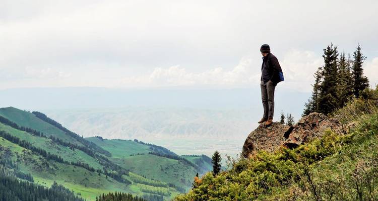 Mann steht auf einer Klippe und blickt auf eine bergige Landschaft.