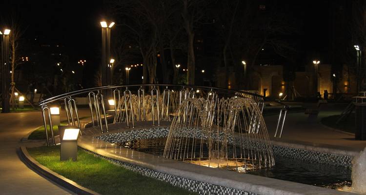 Parc de nuit avec un pont éclairé et une fontaine.