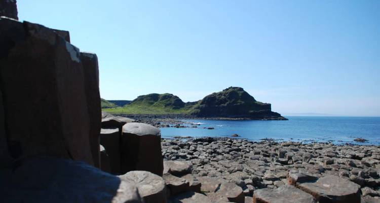 Basaltzuilen en rotsachtige kust van de Giant's Causeway met een grasrijke landtong die tegen een heldere blauwe hemel uitstijgt.