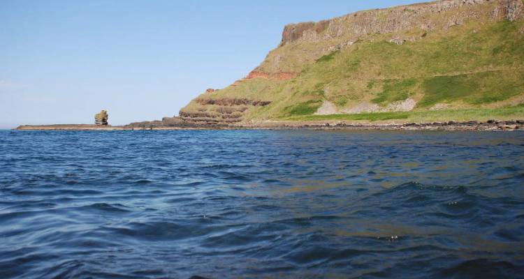 Uitzicht op waterhoogte van een gelaagde kustklif met roestkleurige strepen die een golvende zee ontmoeten.