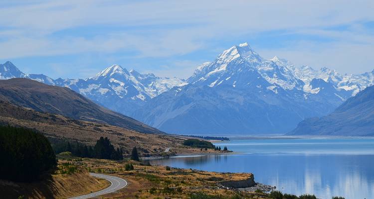 Kronkelende weg langs een meer met majestueuze besneeuwde bergen op de achtergrond.