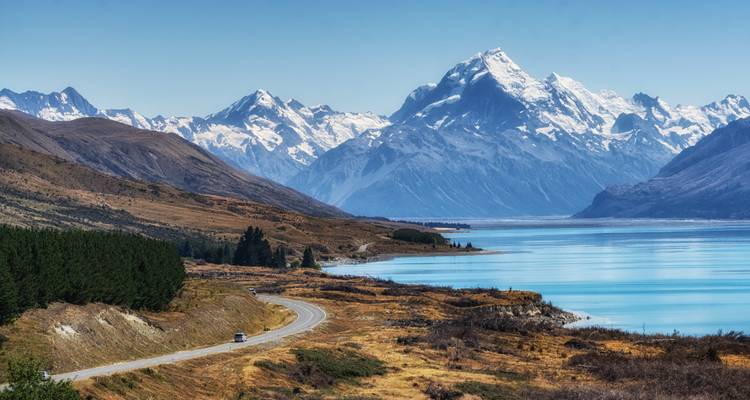 Weidse alpenmeer met turquoise water dat leidt naar een torenhoge sneeuwbedekte piek voorbij glooiende heuvels en kronkelende weg