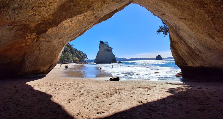 Ein malerischer Blick auf einen Strand durch einen großen natürlichen Felsbogen, mit Menschen, die auf dem Sand wandeln und Wellen, die am Ufer brechen.