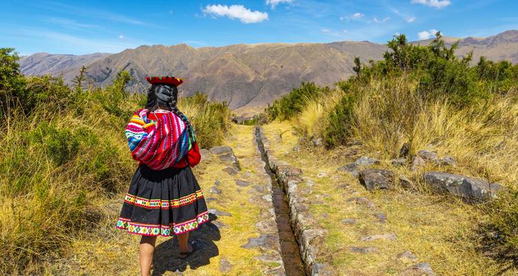 Una mujer en traje tradicional peruano camina por un antiguo canal de riego de piedra con colinas andinas enfrente.