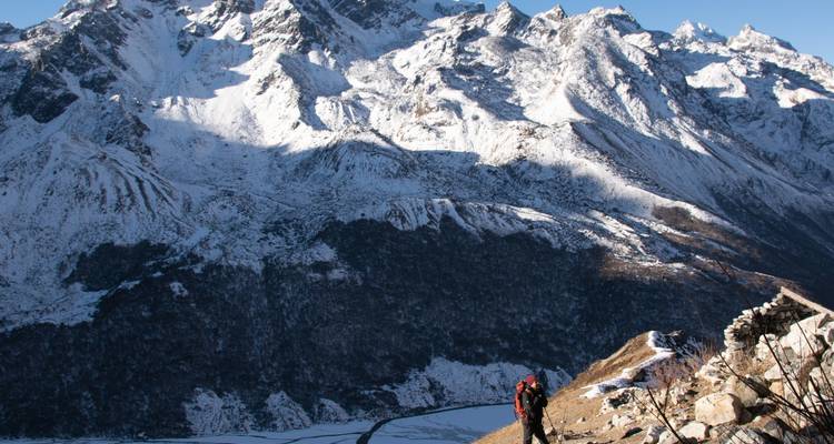 Un randonneur debout sur une pente de montagne avec des pics enneigés en arrière-plan.