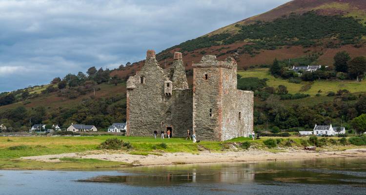 Historische Burgruinen mit Spaziergängern in der Nähe, vor einer Berglandschaft.