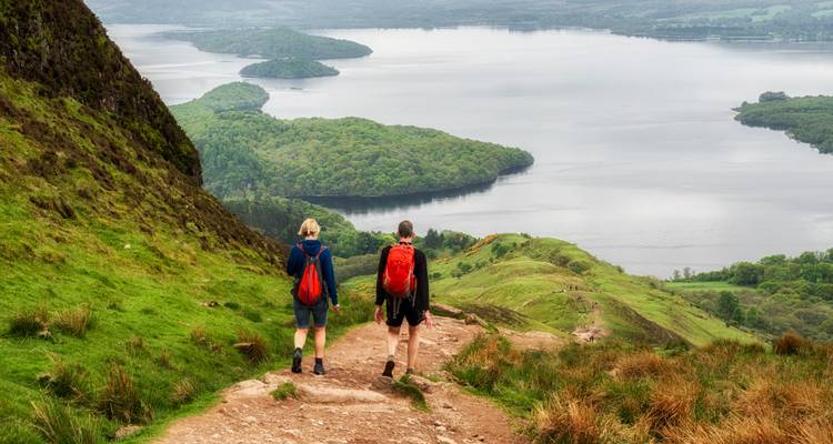 Zwei Wanderer auf einem Bergpfad mit Blick auf einen See und eine üppige Landschaft.
