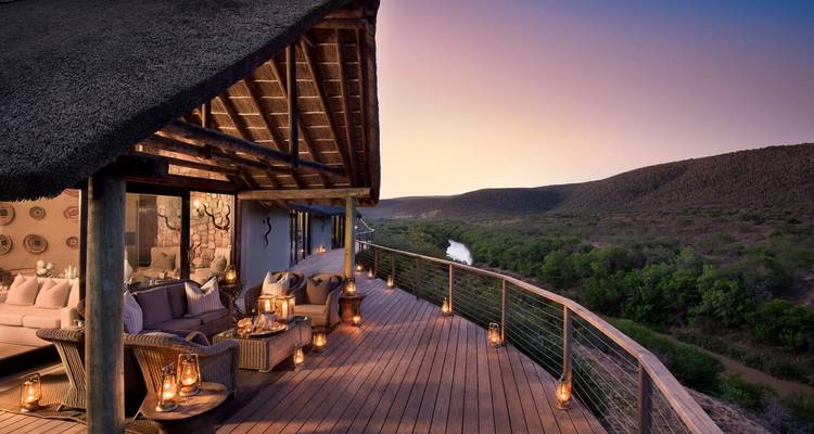 Terrasse extérieure avec sièges confortables et lanternes donnant sur un paysage fluvial pittoresque au coucher du soleil.