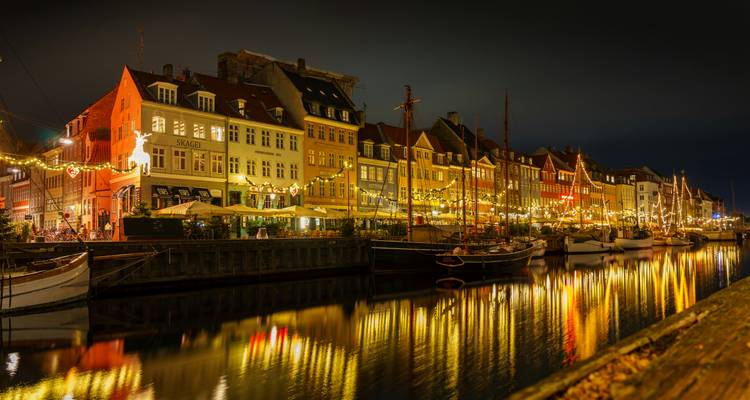 Ein lebendiger Hafenbereich mit gut beleuchteten Gebäuden und Booten, die sich nachts im Wasser spiegeln.
