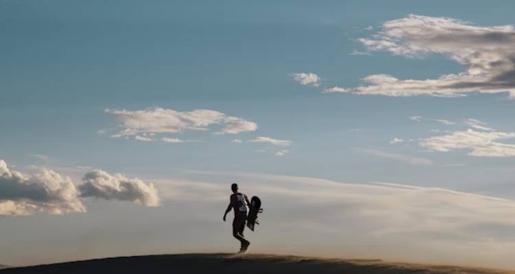 Un hombre caminando con una tabla sobre una duna de arena bajo un cielo vasto.