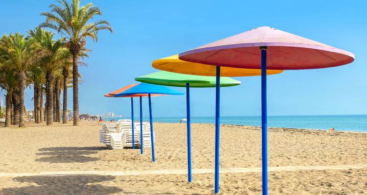 Des parasols colorés en forme de parapluie et des rangées ordonnées de palmiers bordent une plage de sable méditerranéenne vide.