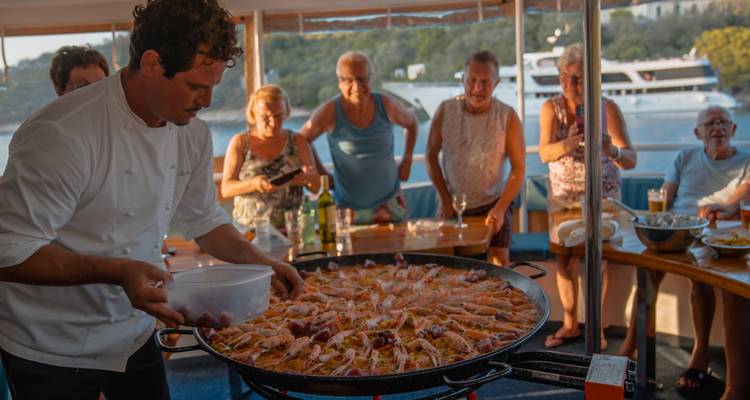 Chef présentant un grand plat de paëlla, des personnes debout en arrière-plan.