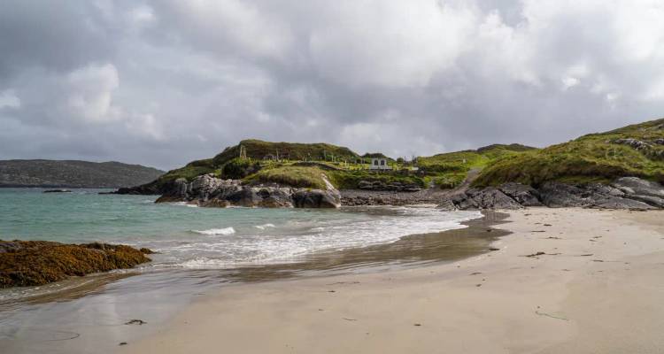 Quiet sandy beach with turquoise waves, rocky headland and distant islands under a cloudy sky.