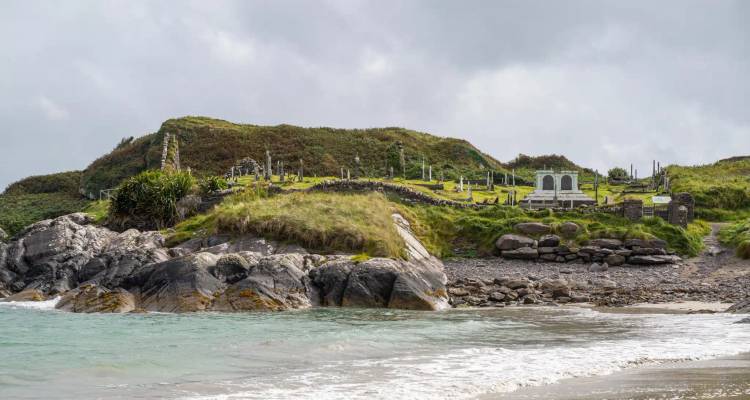 Rocky shoreline with gentle surf and a grassy hill crowned by an old cemetery overlooking the sea.