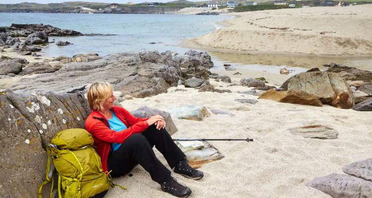 Female hiker rests on a sandy, rocky beach beside her backpack looking out to the sea.