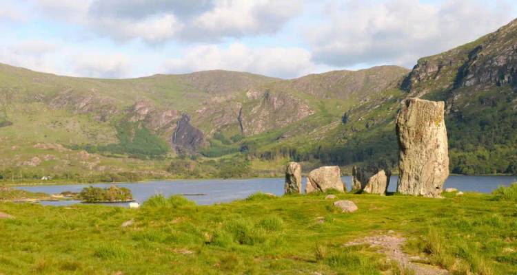 Ancient standing stones overlook a tranquil lake backed by green hills.