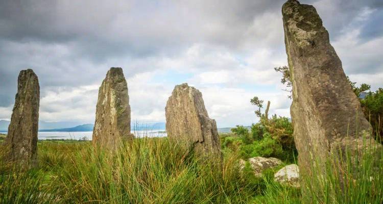 Tall weathered standing stones rise from long grass with a lake and mountains in the distance.