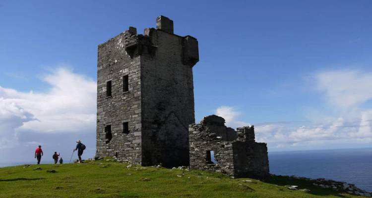 Hikers approach a weathered stone tower ruin perched on a grassy cliff above the Atlantic.