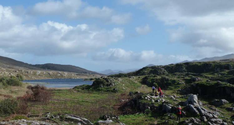 Small group of walkers traverse a rocky green landscape beside a lake under scattered clouds.