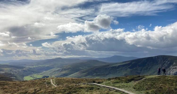 Sweeping vista over the Wicklow Mountains with a boardwalk trail and two hikers pausing on a ridge beneath dramatic clouds.