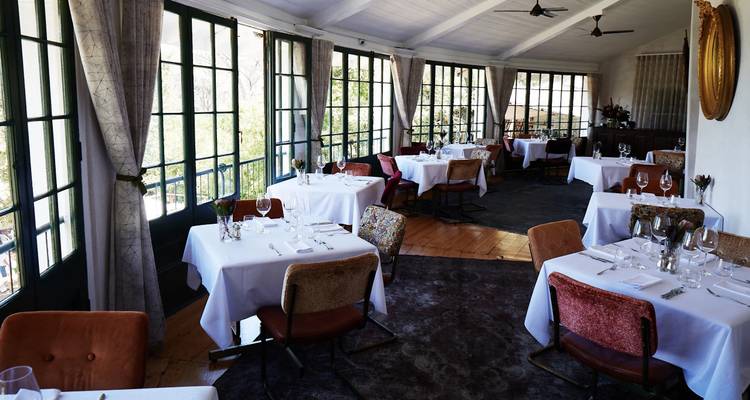 Innenrestaurant mit Tischen und Stühlen vor großen Fenstern.