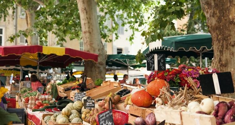 Buitenboeren markt onder bladrijke platanen met kleurrijke producten uitgestald in houten kratten.