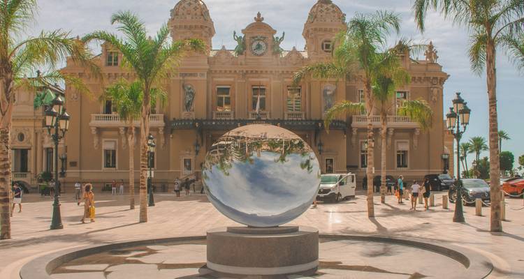 Reflecterende metalen bolsculptuur voor het sierlijke Monte Carlo Casino omringd door palmbomen.