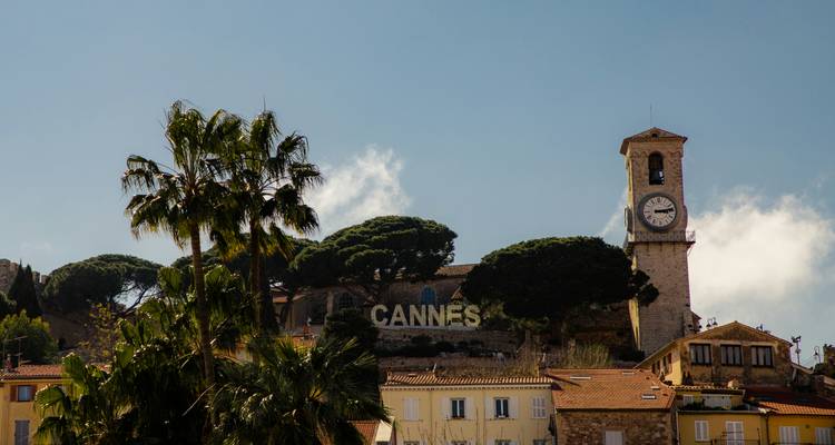 Heuveltopzicht op Cannes met klokkentoren en groot 'CANNES' bord omlijst door palmbomen.