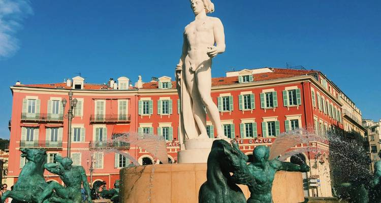 Fontein met klassiek naakt standbeeld omringd door bronzen figuren voor kleurrijke historische gebouwen onder een heldere blauwe hemel