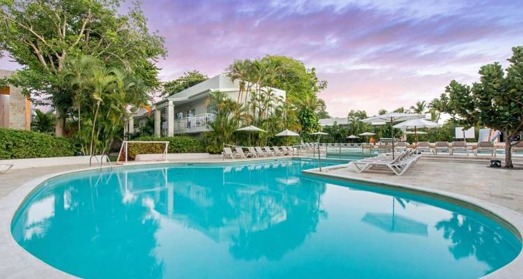 A poolside view with a cushioned seating area, palm trees, and a sunset sky.