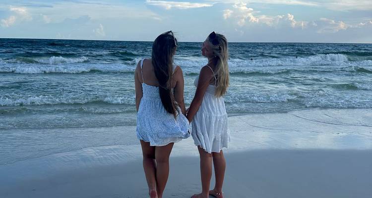 Two people in white dresses on a beach looking at the ocean.