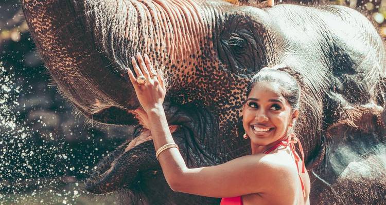Woman smiling while touching an elephant with water splashing around.