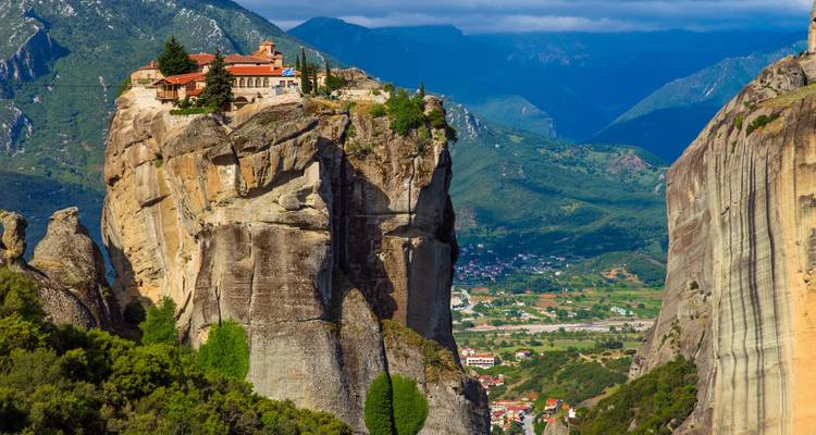 Monasterio encaramado dramáticamente en la cima de un pilar de arenisca imponente con un valle exuberante y montañas detrás.