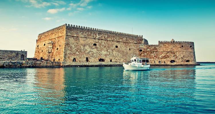 Fortaleza marítima medieval de piedra con murallas almenadas reflejándose en aguas azules tranquilas con un pequeño bote blanco.