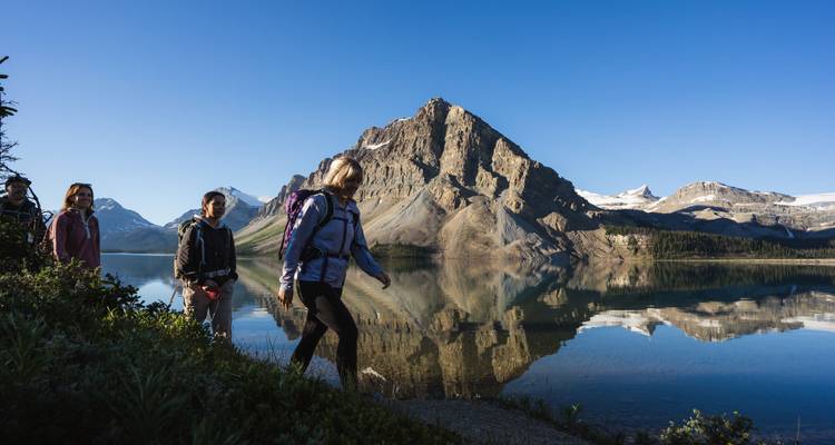 Excursionistas junto a un lago con el reflejo de una montaña en un cielo azul claro.
