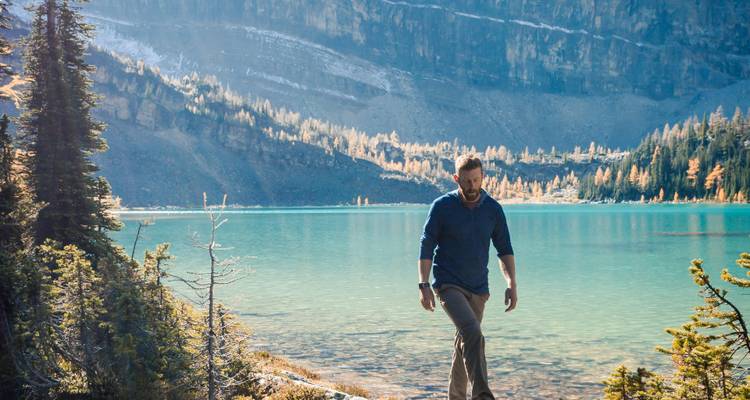 Un hombre caminando junto a un lago turquesa con bosque al fondo.