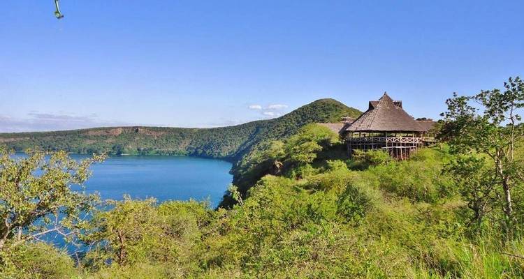 Lodge aux toits de chaume perché sur une falaise verdoyante surplombant un lac de cratère bleu turquoise