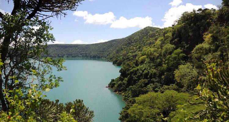 Des pentes forestières verdoyantes descendant vers un lac de cratère bleu-vert sous un ciel dégagé