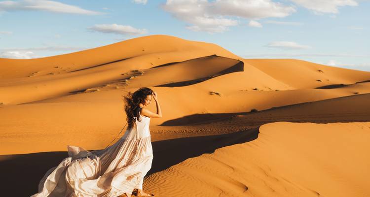 Een vrouw in een wapperende jurk die bij zonsondergang over zandduinen loopt.