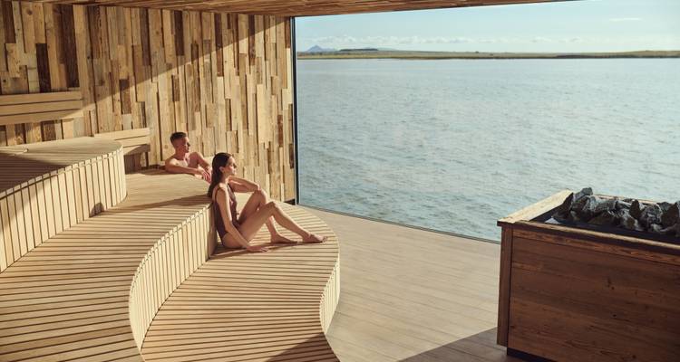 Dos personas relajándose en una sauna de madera con vista al agua.