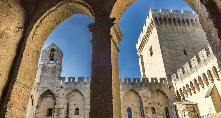 Arches de pierre encadrant les murs fortifiés et la tour d'un palais médiéval contre un ciel bleu clair.