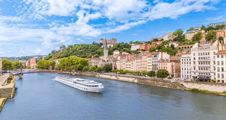 Bateau de croisière fluviale glissant devant des bâtiments colorés au bord de la rivière et des collines couvertes d'arbres sous un ciel bleu éclatant.