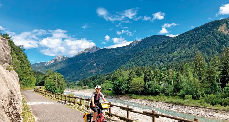 Einzelner Radfahrer genießt einen Radweg im Alpental mit klarem Fluss und bewaldeten Bergen unter blauem Himmel.