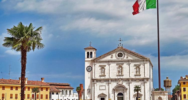 Historische Marmorkirche auf italienischem Platz mit wehender Flagge und Palme unter strahlend blauem Himmel.
